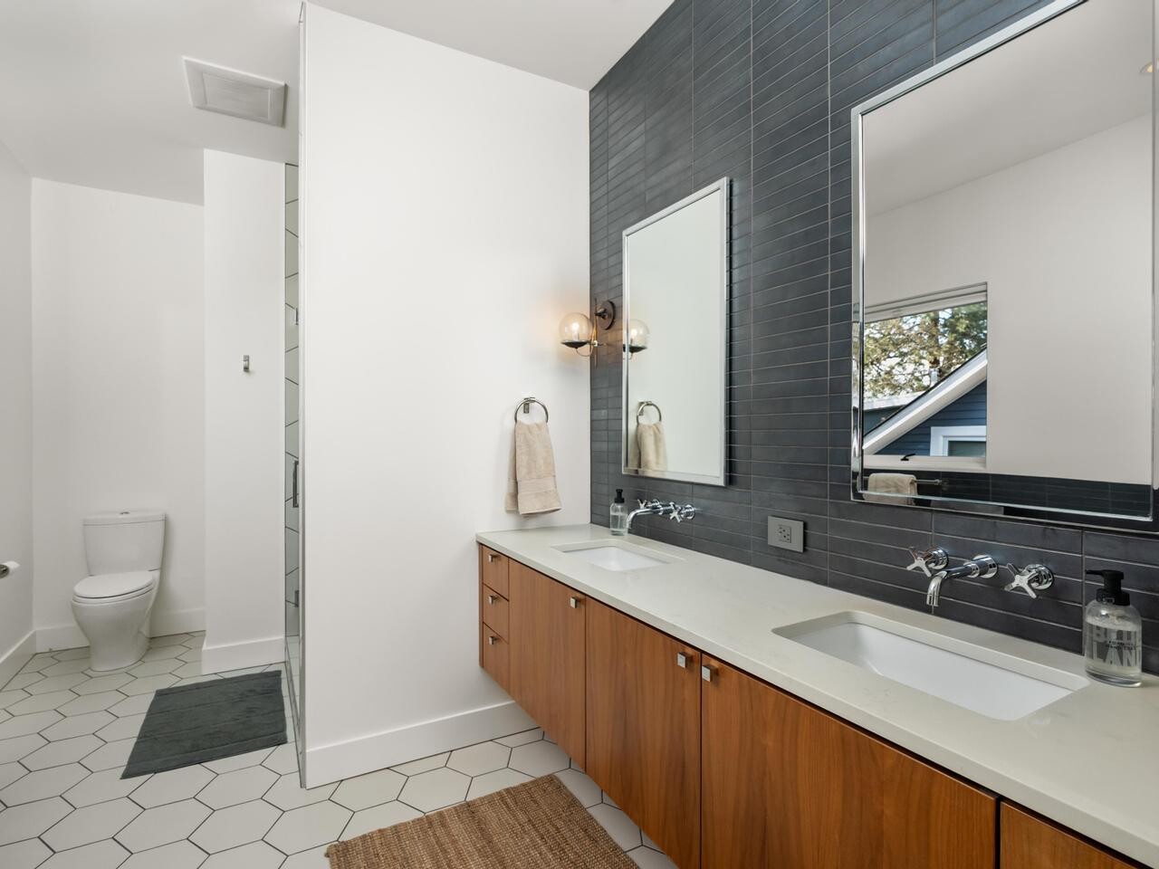 Modern bathroom with a double sink vanity, wooden cabinets, two mirrors, black tile accent wall, hexagon floor tiles, and a separate area for the toilet. A towel and soap dispenser are on the vanity.