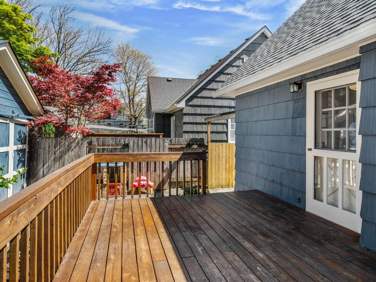 A wooden deck with a railing attached to a blue house, overlooking a backyard with trees, a fence, and neighboring houses; a door with glass panels leads inside.