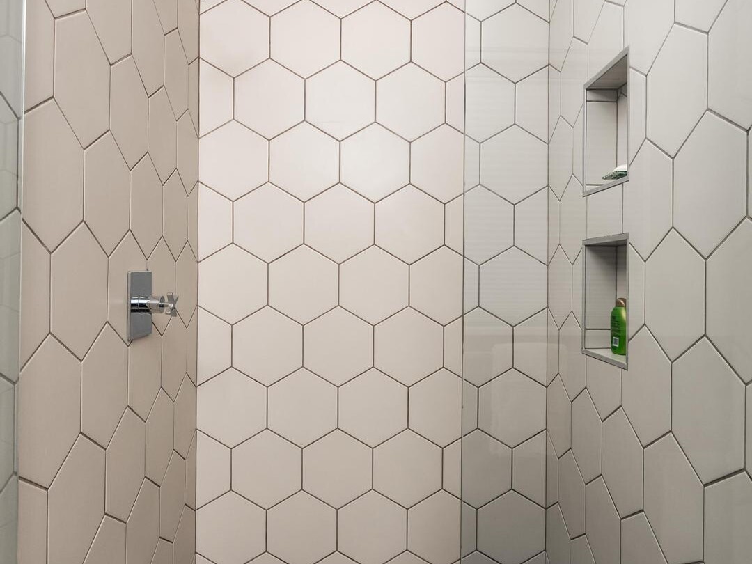 A modern shower with large beige hexagonal wall tiles, a rainfall showerhead, built-in shelves holding toiletries, and a glass partition. The floor features small black hexagonal tiles and a central square drain.