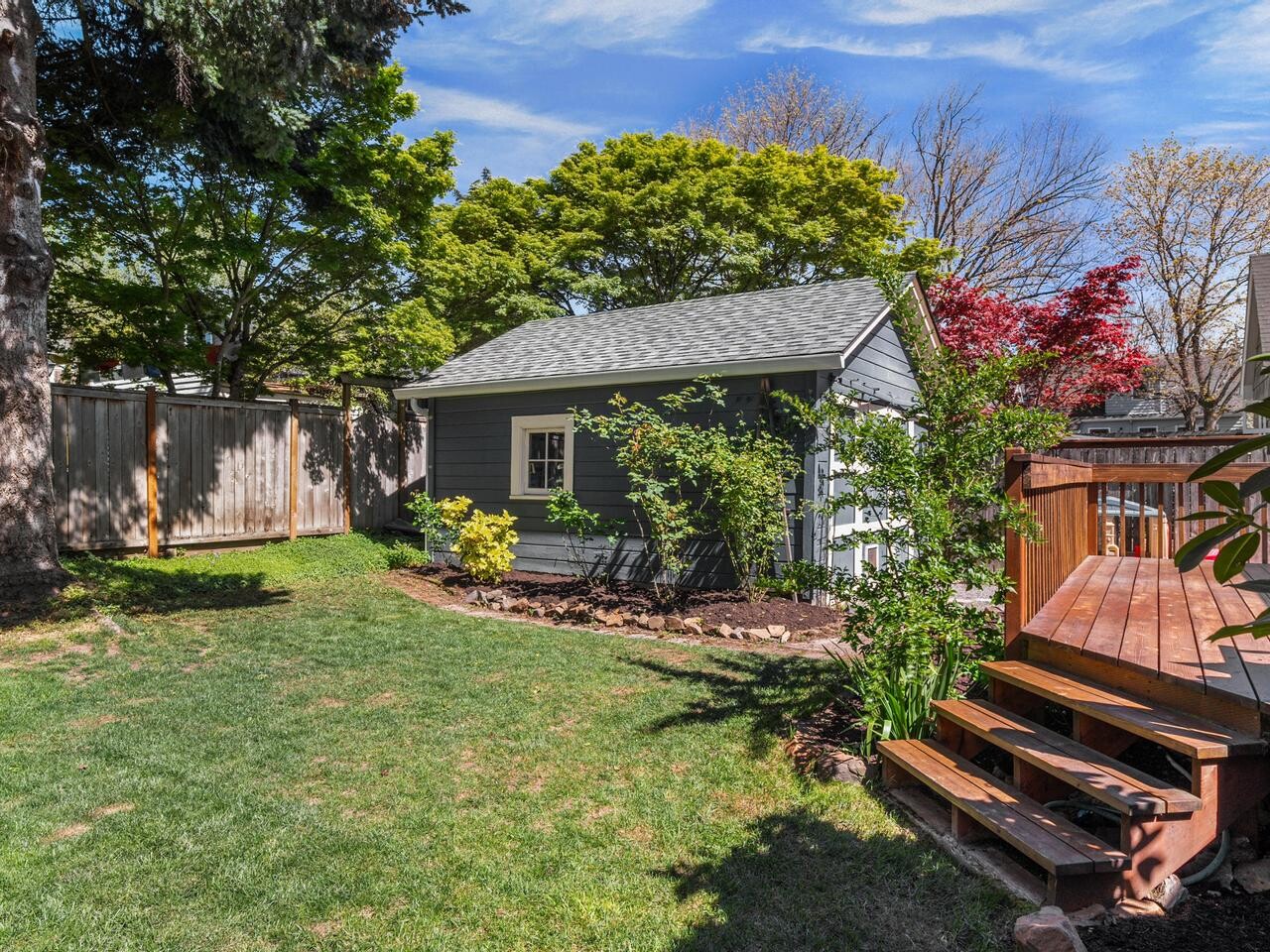A small gray shed with white trim sits in a backyard with green grass, mature trees, and a wooden fence. Wooden steps lead up to a deck on the right, with lush plants and spring foliage throughout the scene.