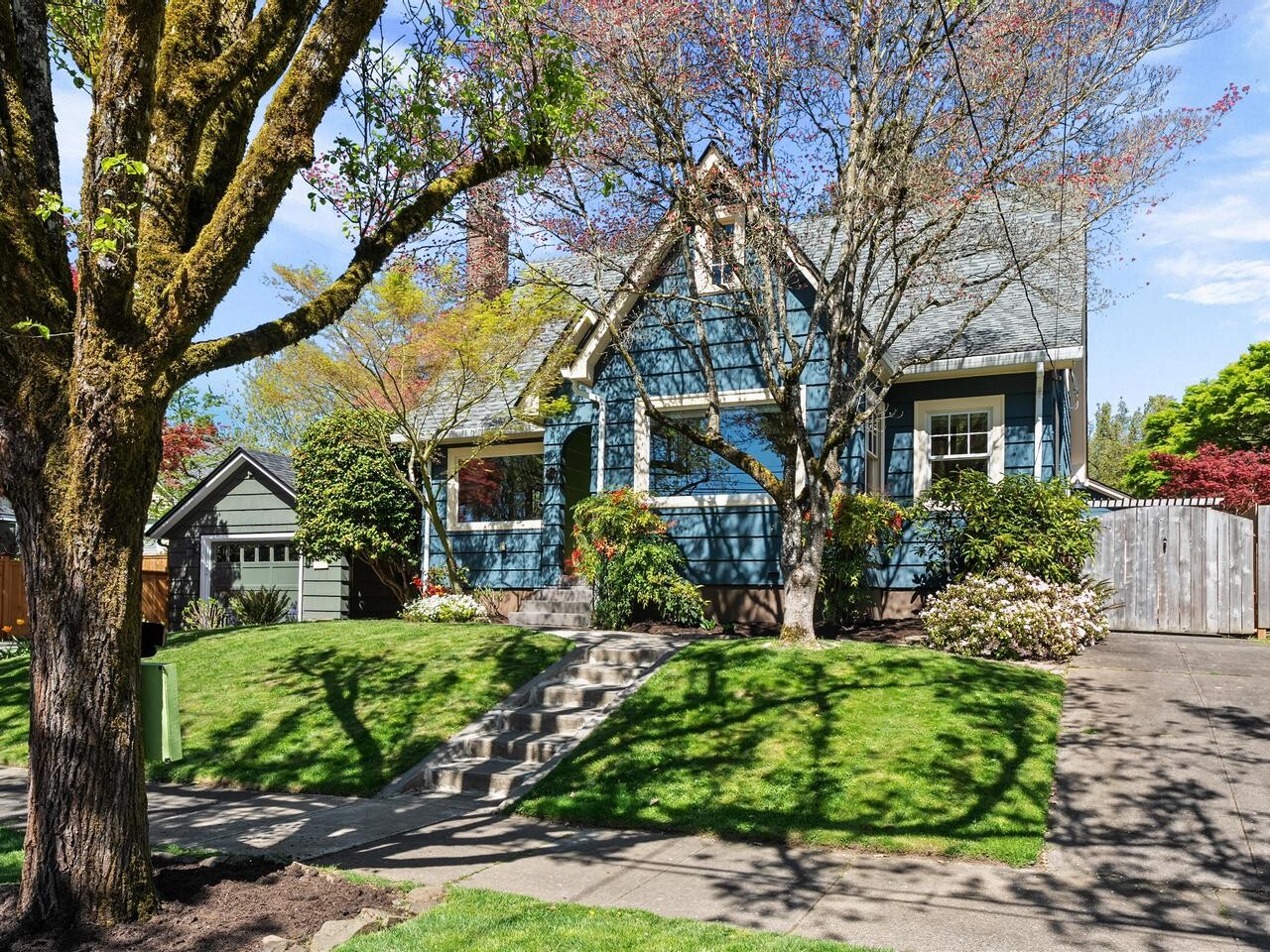 A charming blue house with white trim sits behind a well-kept lawn and trees, featuring a front porch, steps leading up to the entrance, and a detached garage to the left.