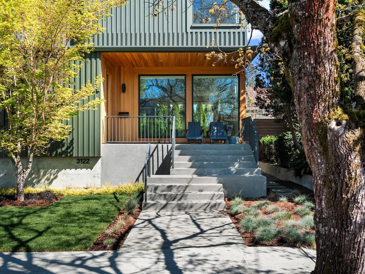 Modern house with green siding, large windows, and a wooden overhang above a concrete porch. Steps lead up from the sidewalk, with trees and landscaped greenery in the front yard. House number 3122 is visible.