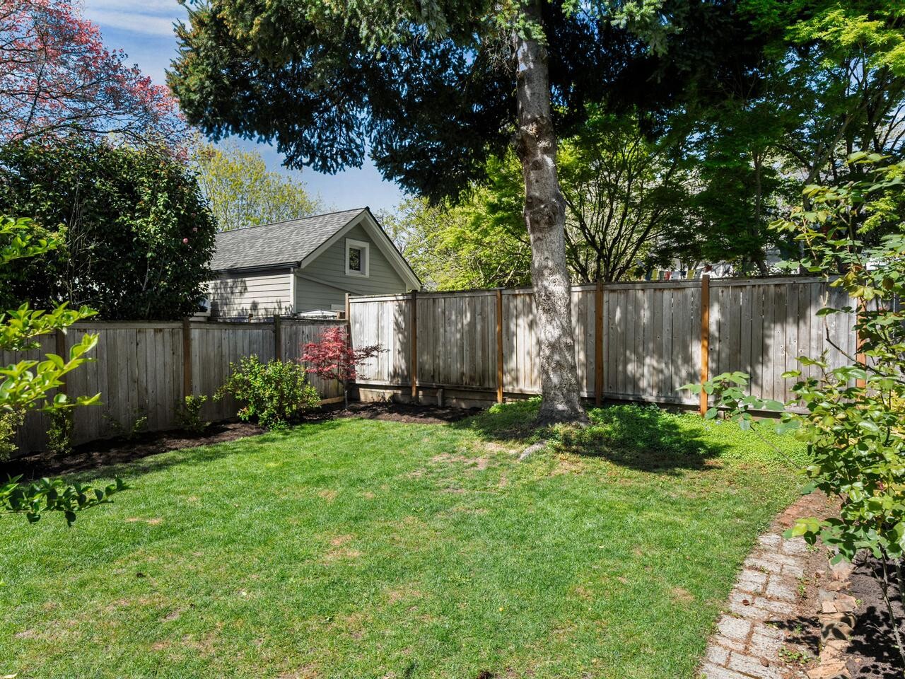 A small backyard with green grass, surrounded by a wooden fence, trees, and bushes. A brick path curves along one edge, and a gray house with a sloped roof is visible beyond the fence.