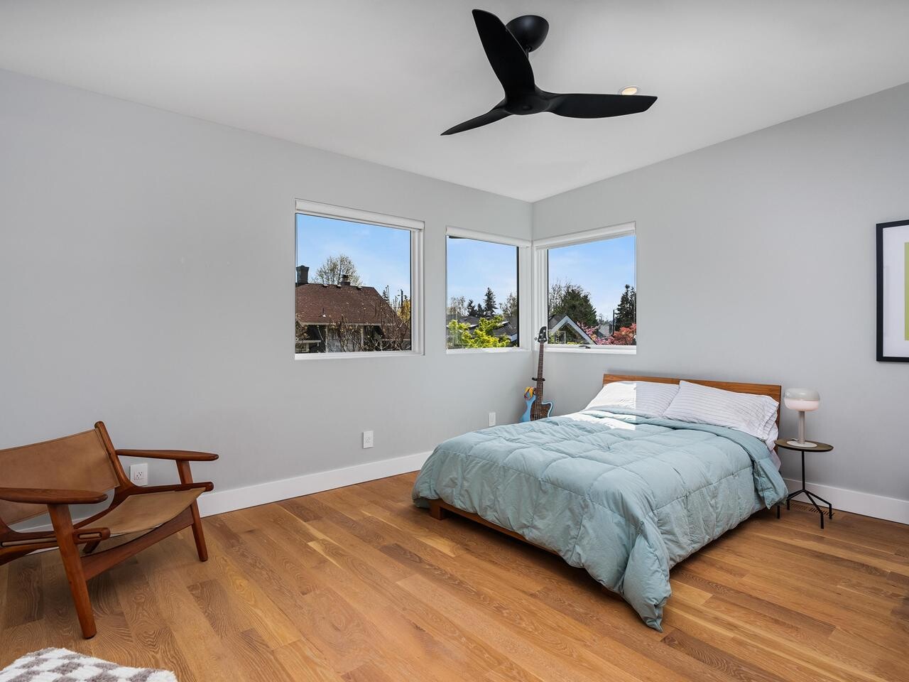 Minimalist bedroom with light gray walls, hardwood floor, a large bed with a light blue comforter, two windows with outdoor views, a modern ceiling fan, wooden chair, small side table, and framed artwork on the wall.