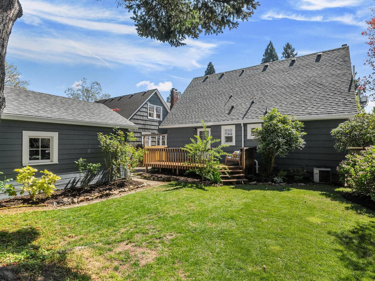 A well-maintained backyard with green grass, garden beds, and shrubs. A wooden deck with steps leads to the back door of a gray house with a steep roof, next to a detached garage, under a partly cloudy sky.
