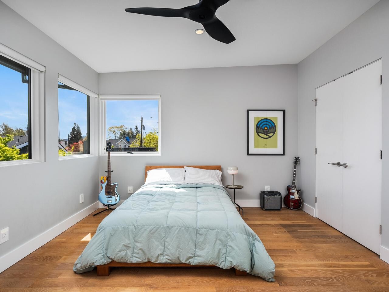 Modern bedroom with a single bed, pale blue comforter, two windows with outdoor views, a ceiling fan, two guitars, a framed poster, and wooden floors. Room is minimalistic with white walls and natural light.