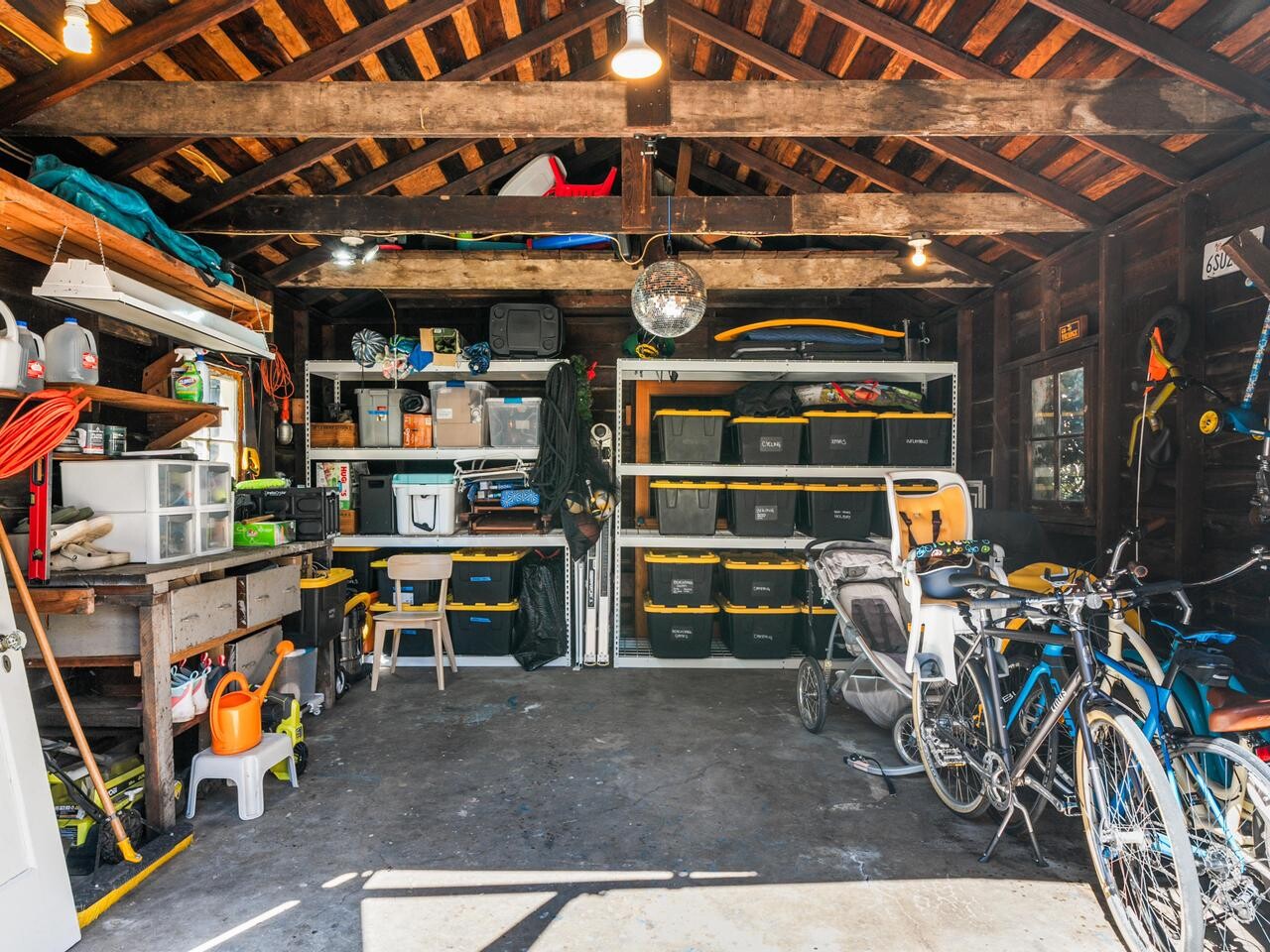 A well-organized garage with shelves holding storage bins, tools, and equipment. On the right, several bicycles are parked. The space has exposed wooden beams and is well-lit by overhead lamps.