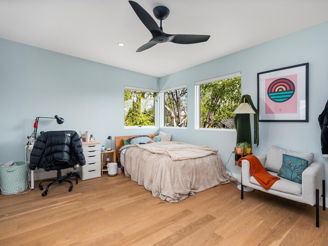 A cozy bedroom with light blue walls, wooden floor, a bed with beige bedding, a small desk and office chair, a white armchair, two large windows with trees outside, and modern decor including framed art and a floor lamp.