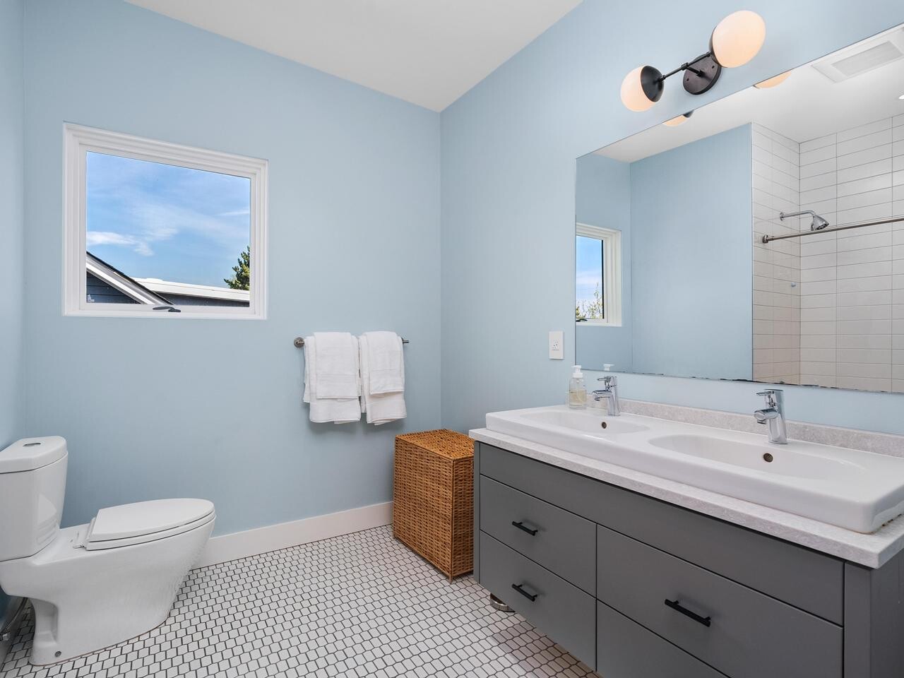 Modern bathroom with light blue walls, a white toilet, double-sink gray vanity, large mirror, hexagon tile floor, wicker laundry basket, and two white towels hanging by a window. A shower area is visible in the background.