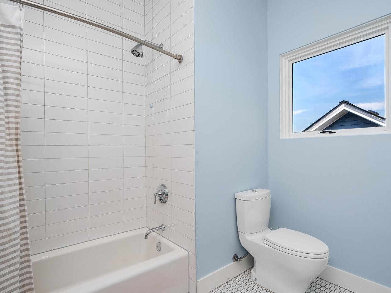 A clean, modern bathroom with a white bathtub and striped shower curtain on the left, white subway tile walls, a white toilet on the right, and a window with a view of a blue sky and rooftops.