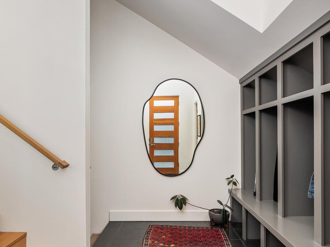 A modern entryway with a wavy-edged mirror above a red patterned rug, gray cubby storage on the right, wooden stairs on the left, and a potted plant near the mirror. The area features white walls and black tile flooring.