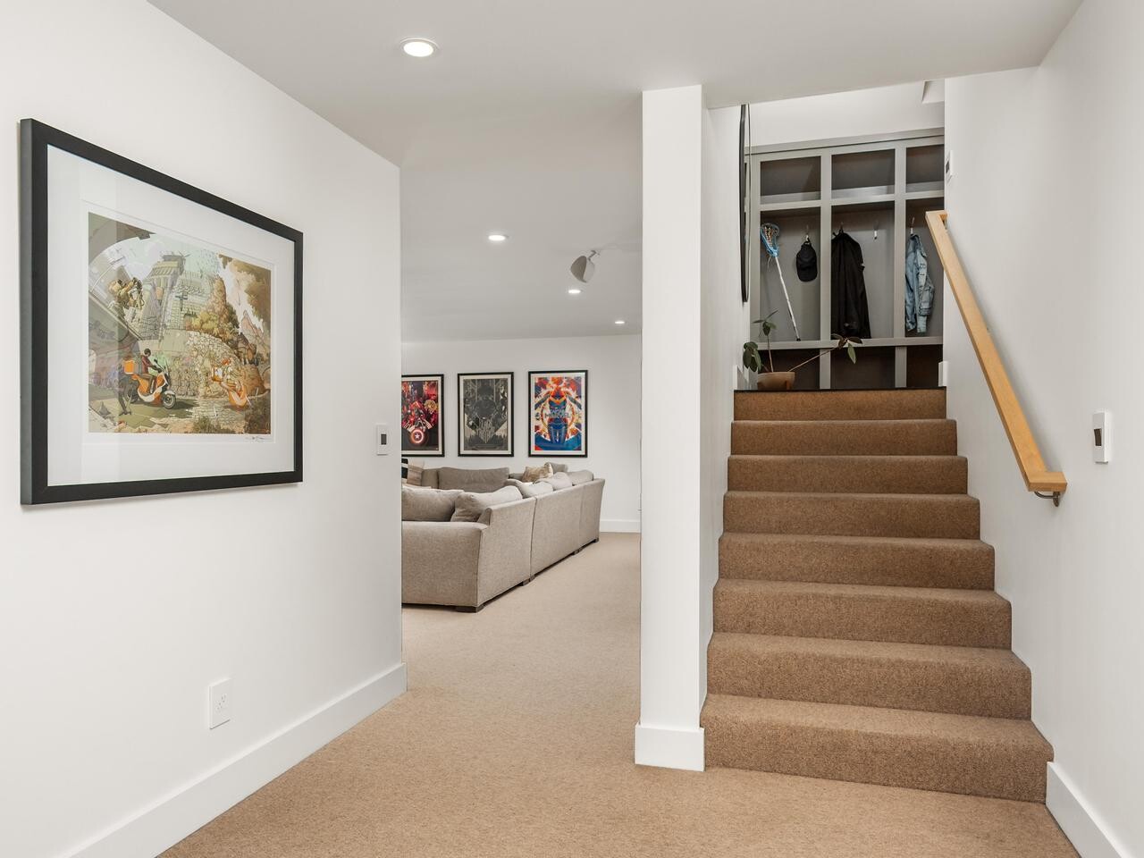 A carpeted staircase with a wooden handrail leads down to a modern basement living area with beige carpet, neutral-colored sofas, and framed artwork on white walls. A coat rack with hanging clothes sits near the stairs.