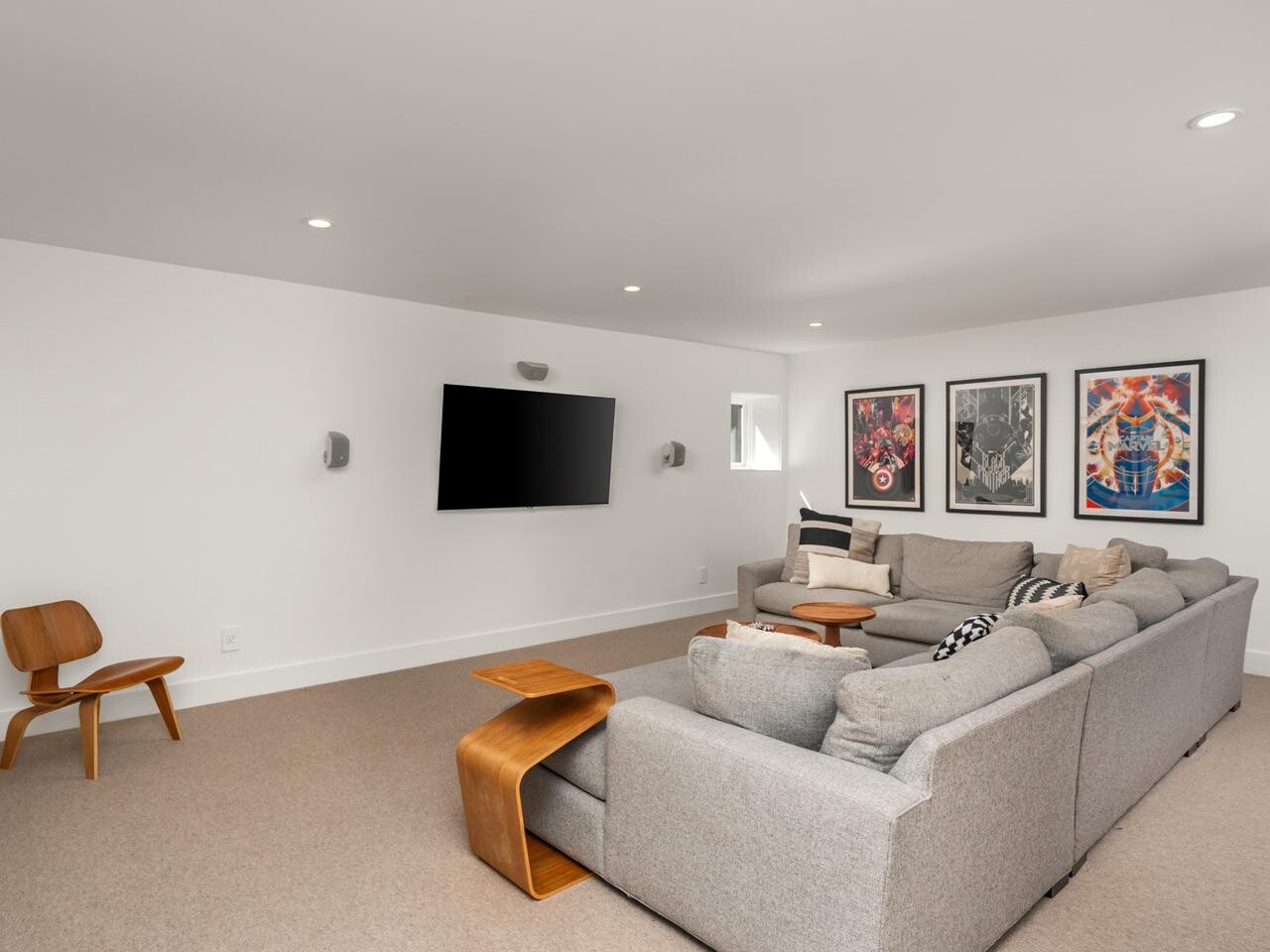 A modern home theater room with a large gray sectional sofa, a wall-mounted flat-screen TV, a wooden chair, and three colorful framed art pieces on the white walls. The space is well-lit with recessed ceiling lights.