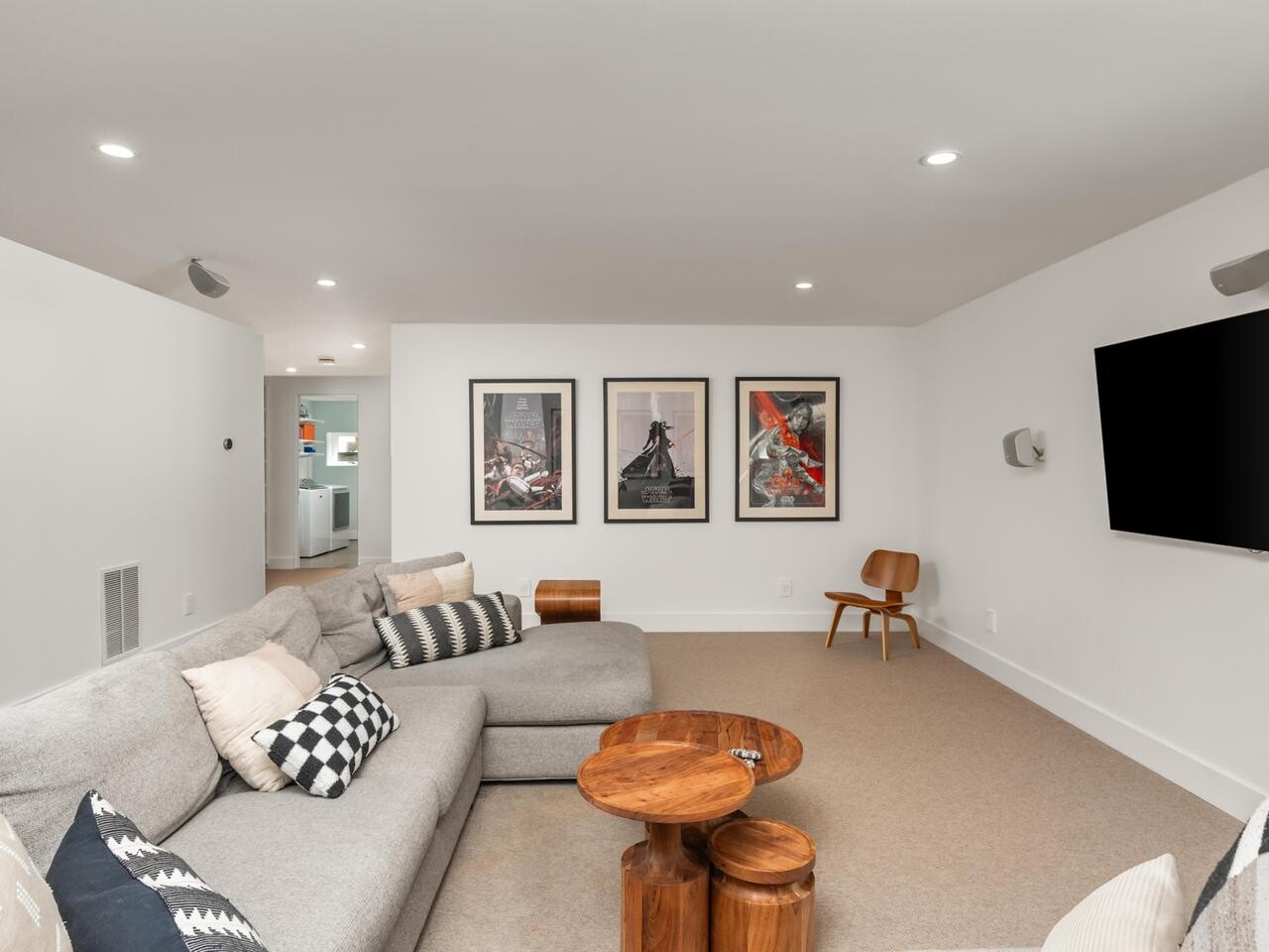 A modern living room with a large beige sectional sofa, patterned pillows, round wooden coffee tables, framed artwork on a white wall, a wooden chair, and a flat-screen TV mounted on the opposite wall.