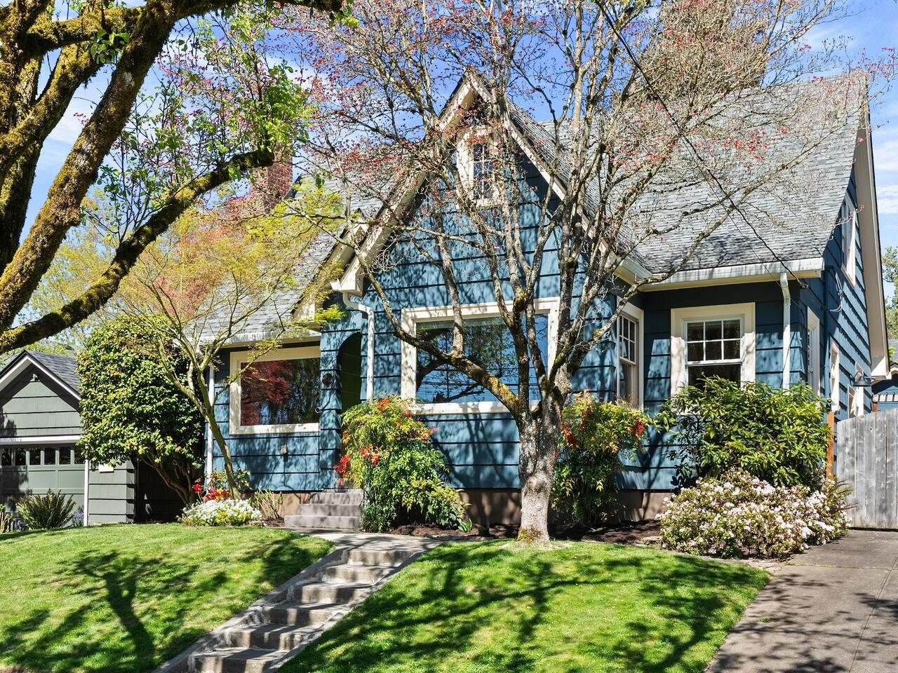 A blue house with white trim, large front windows, and a sloped roof sits behind a well-manicured lawn and flowering shrubs. Trees with sparse leaves shade the yard and sidewalk. A driveway leads to a detached garage on the left.