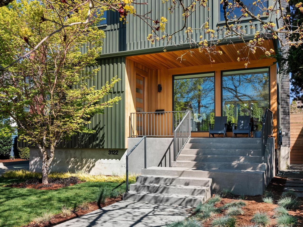 A modern two-story house with green siding, large windows, and a wooden front porch. Concrete steps with a metal railing lead to the entrance, surrounded by trees, grass, and landscaping.