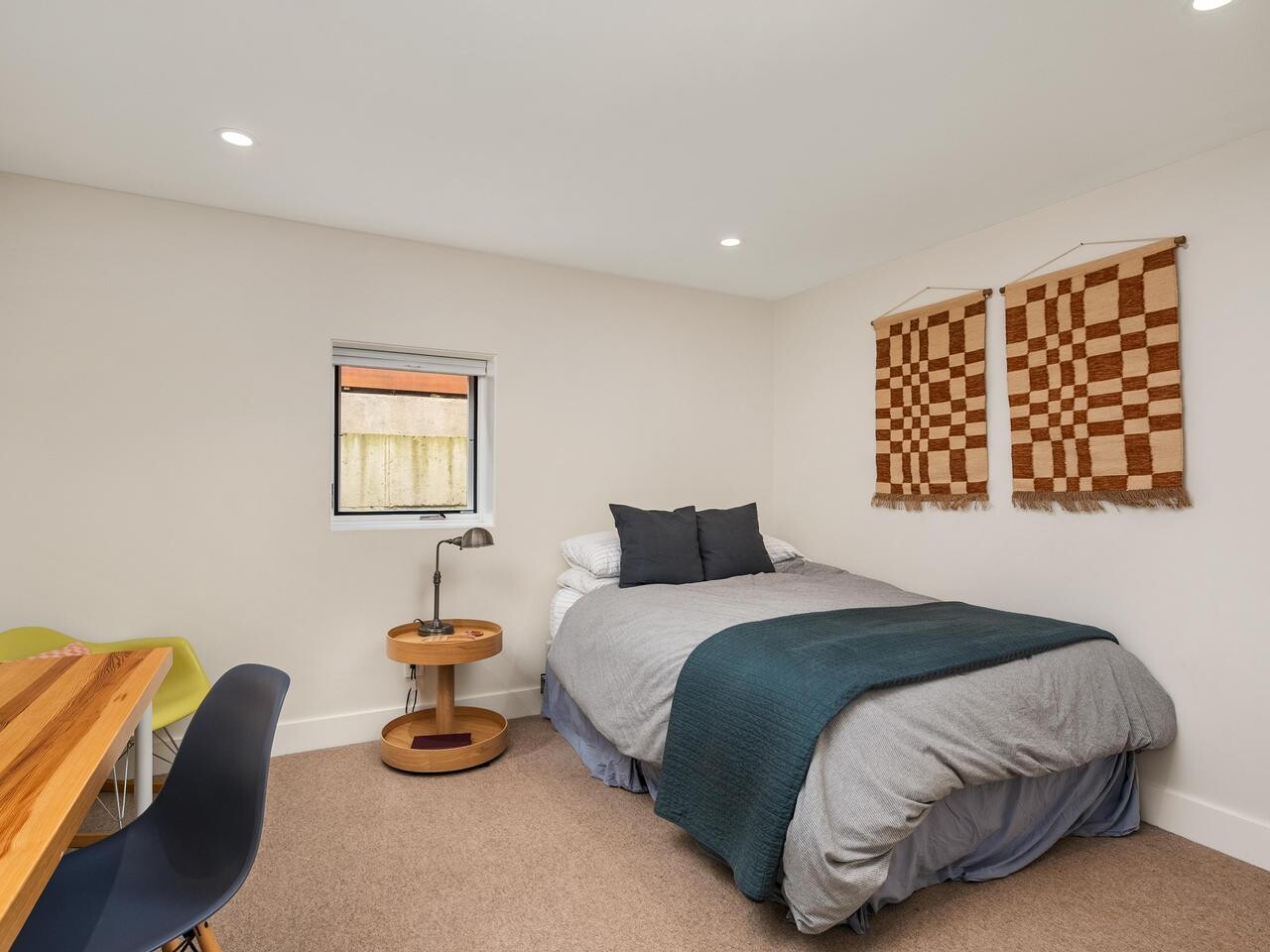 A simple bedroom with a double bed, gray and teal bedding, two checkered wall hangings, a wooden side table with a lamp, a small window, and a wooden desk with two chairs along the left wall.