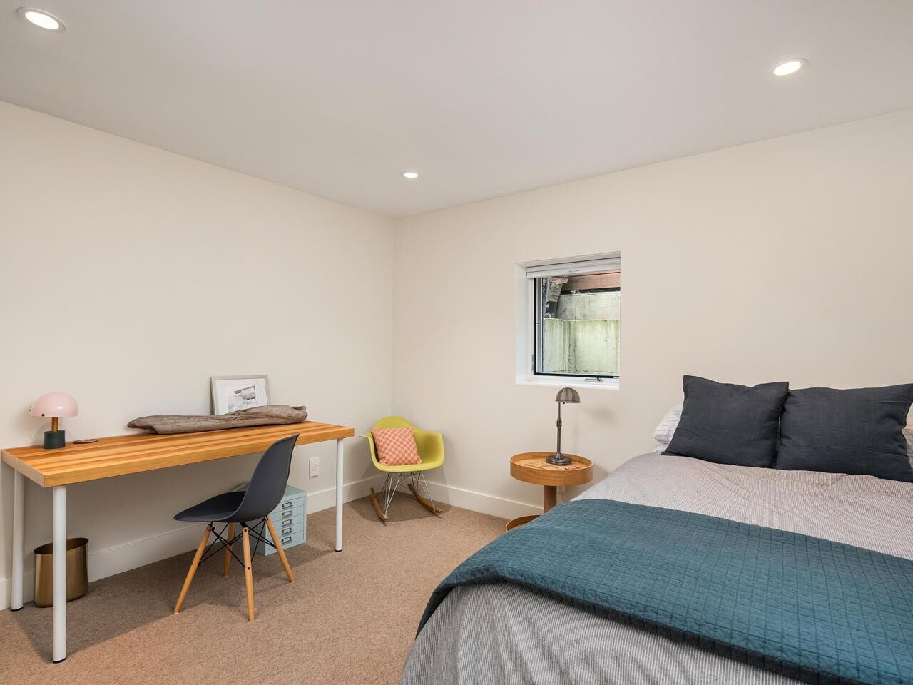A minimalist bedroom with a bed, dark pillows, teal blanket, small round side table with a lamp, a wooden desk with a chair, and a small window letting in natural light.