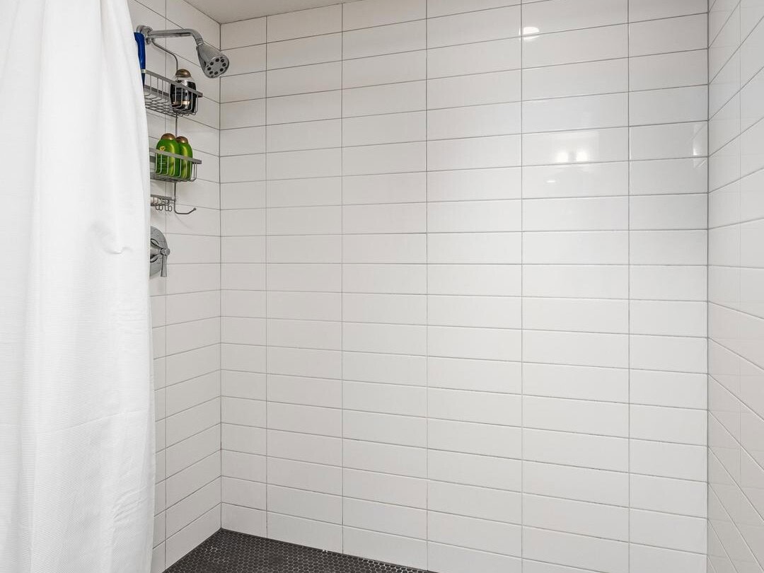 A modern shower with white subway tile walls, a dark textured floor, a white shower curtain, and a metal caddy holding toiletries in the corner. A circular overhead light illuminates the area.