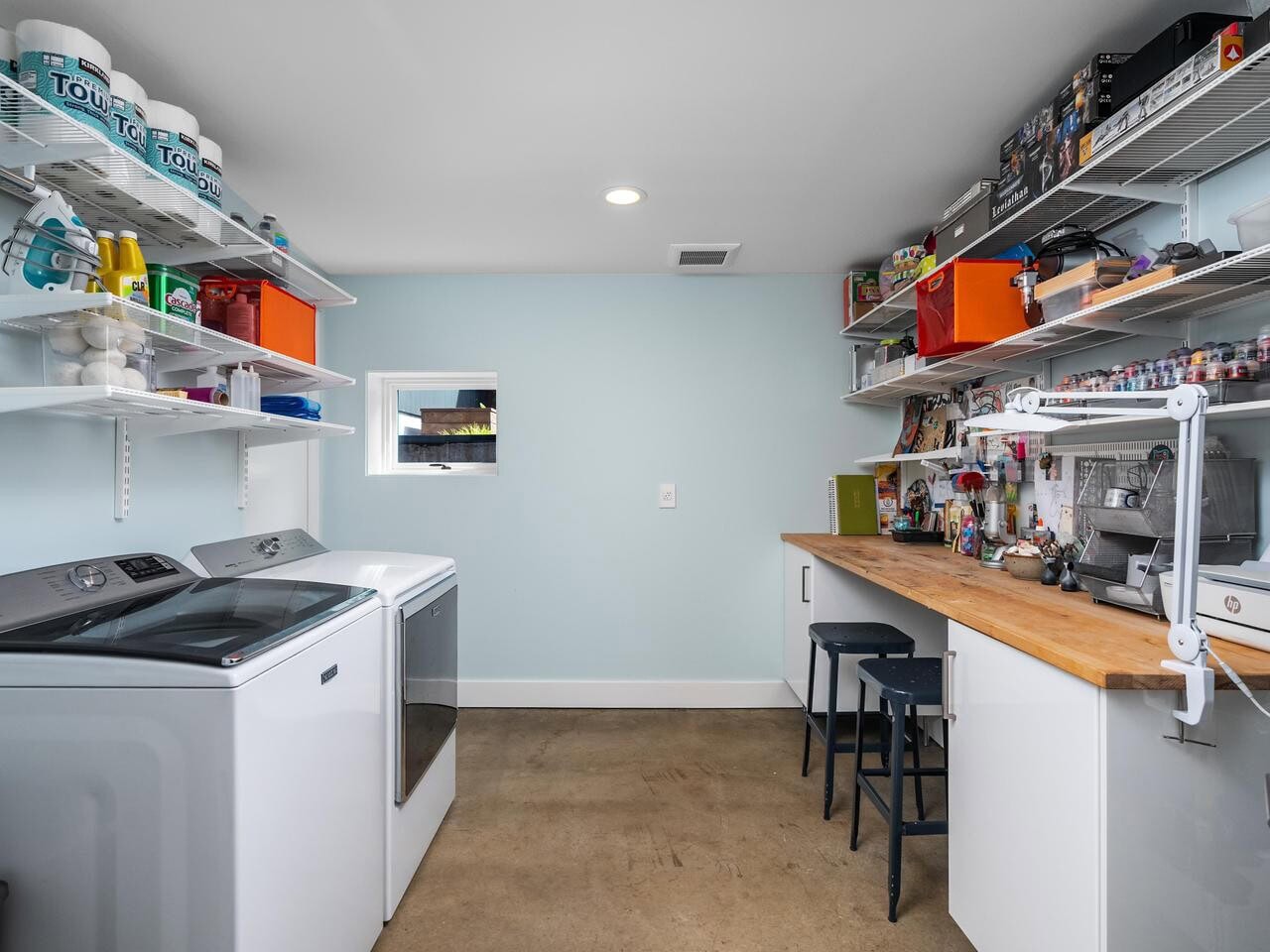 A laundry and craft room with a washer and dryer on the left, shelves holding supplies, a wooden workbench with stools on the right, and a polished concrete floor. The walls are light blue with a small window.