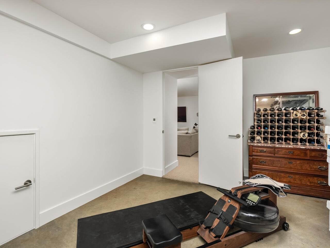 A small room with white walls, a rowing machine on a black mat, a wooden dresser with a wine rack, stacked boxes, and an open door leading to a carpeted living area with a sofa.