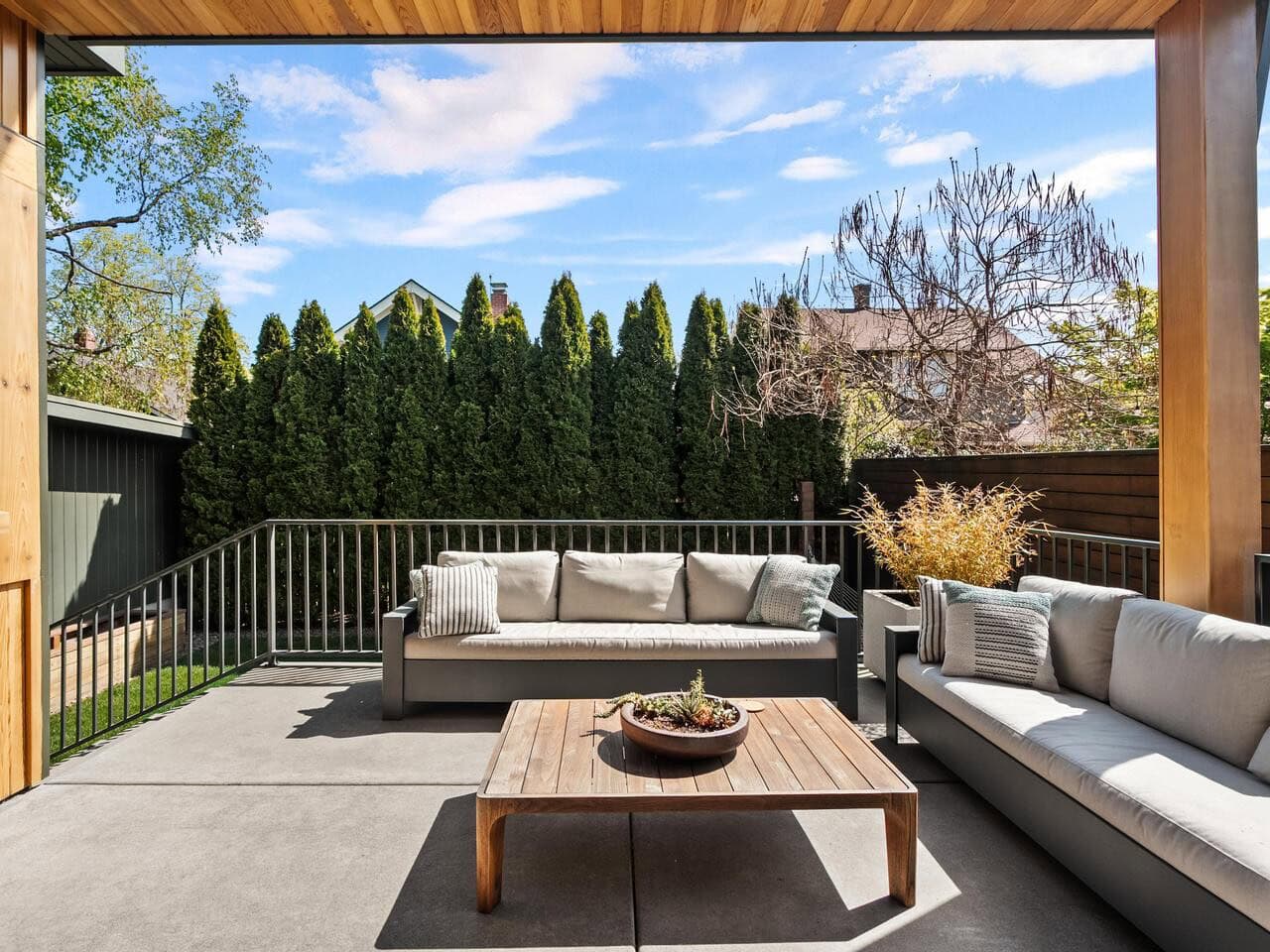 Modern outdoor patio with two gray sofas, a wooden coffee table with a planter, railing, and tall green trees providing privacy. The area is shaded by a wooden overhang and surrounded by greenery under a blue sky.