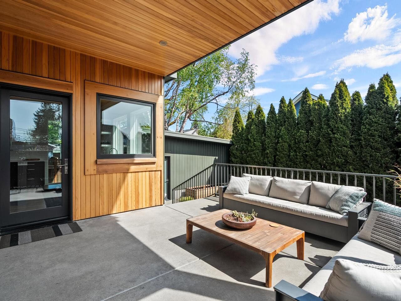 A modern patio with a wood-paneled exterior, a black door, large window, gray outdoor sofas with cushions, a wooden table, and tall green trees along the fence under a blue sky.