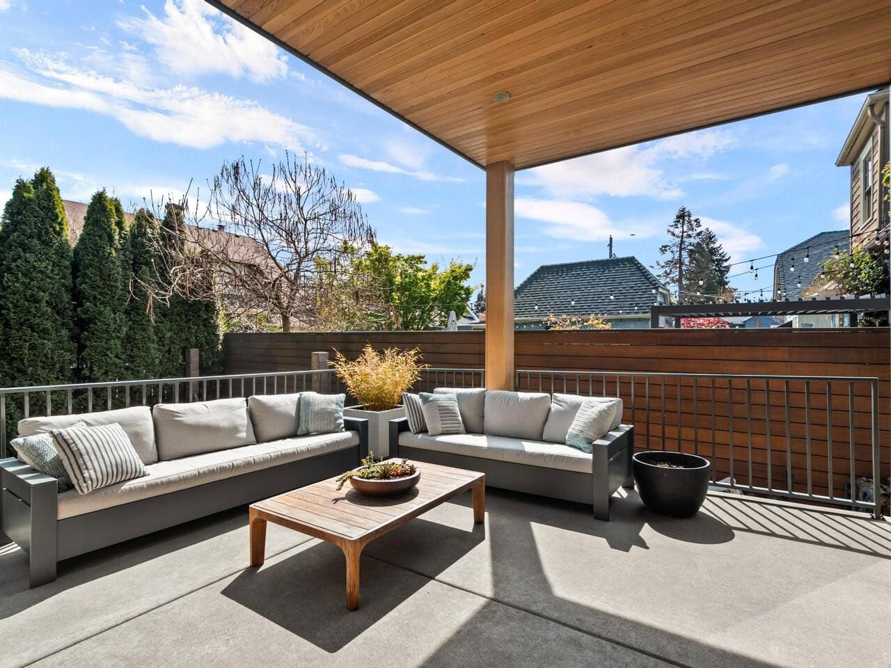 A modern outdoor patio with two gray cushioned sofas, a wooden coffee table, potted plants, and greenery, covered by a wooden ceiling and enclosed by a metal railing and privacy fence.