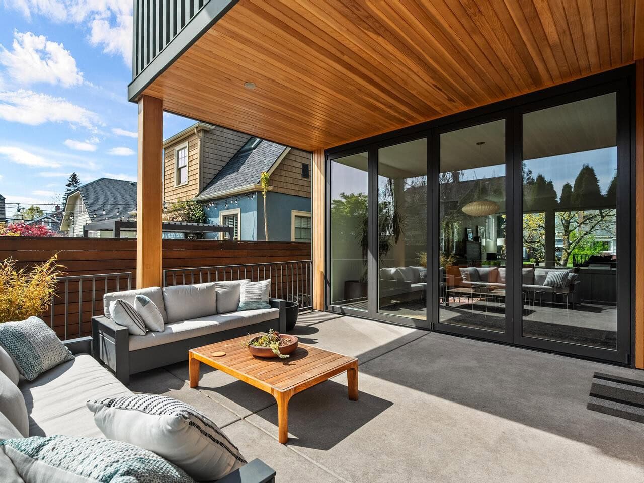 Modern covered patio with wooden ceiling, outdoor sofas, a low coffee table, and large sliding glass doors leading to the interior of a house. The space is bright and surrounded by neighboring homes and greenery.