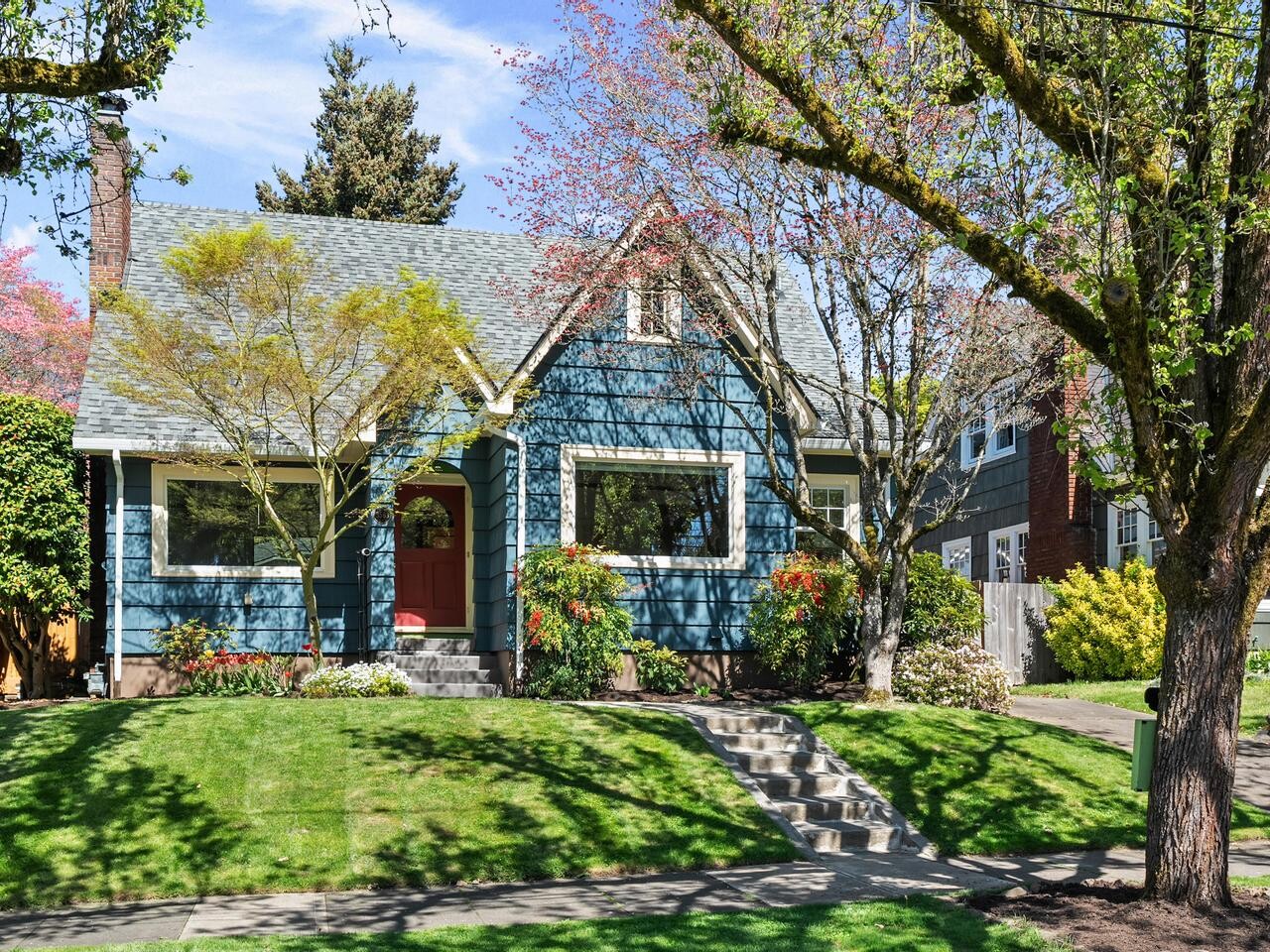 A charming blue house with white trim and a red door sits behind a well-kept lawn and stone steps, surrounded by leafy trees and blooming bushes on a sunny day.