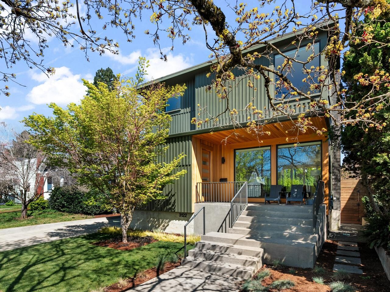 Modern two-story house with green siding and large windows, a covered front porch with seating, a concrete staircase, and a well-maintained lawn with trees and shrubs under a partly cloudy sky.