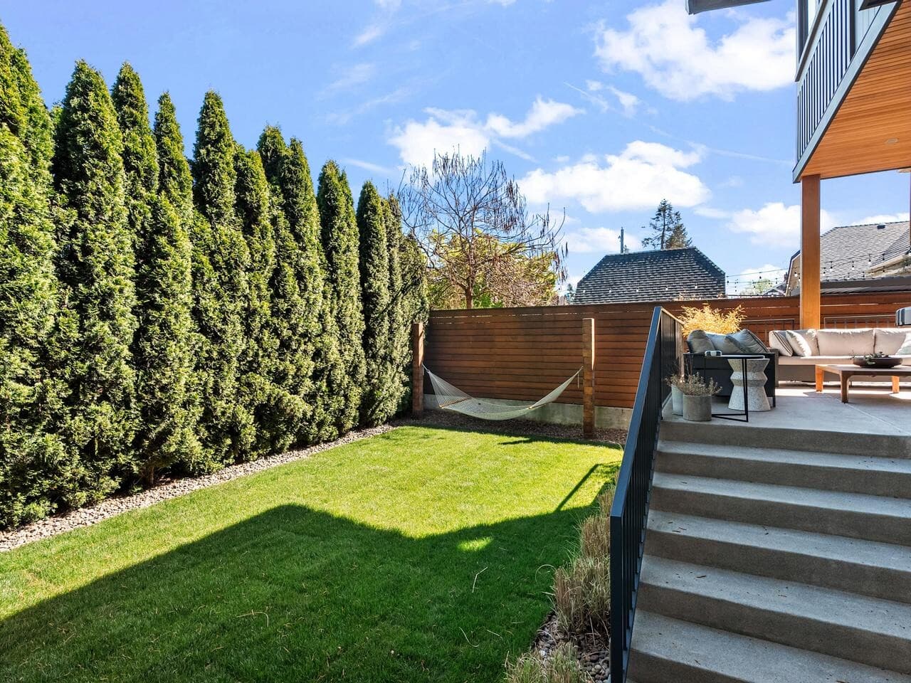 A backyard with a green lawn, tall evergreen trees along the fence, a hammock in one corner, and a patio with outdoor seating at the top of concrete steps. The sky is blue with scattered clouds.