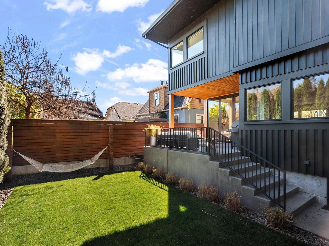 A modern house with dark siding and large windows overlooks a small backyard with green grass, a hammock, wooden fence, and metal-railed stairs leading to a patio area. The sky is blue with scattered clouds.