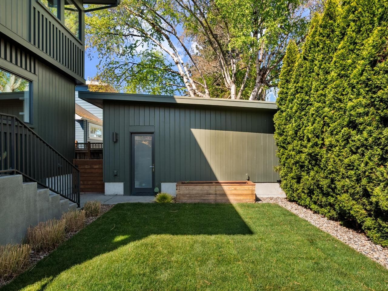 A modern backyard features green siding, a small concrete patio, a raised wooden planter, and a lush lawn bordered by tall evergreen hedges and a row of trimmed plants, with trees and a blue sky in the background.