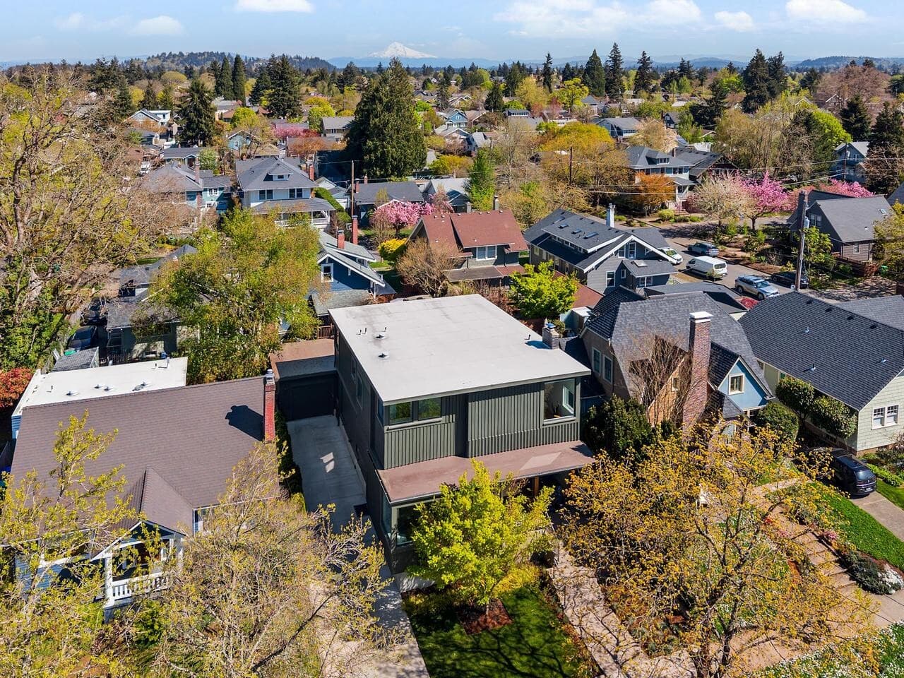Aerial view of a suburban neighborhood with a mix of modern and traditional houses, tree-lined streets, and distant mountains under a blue sky with scattered clouds.