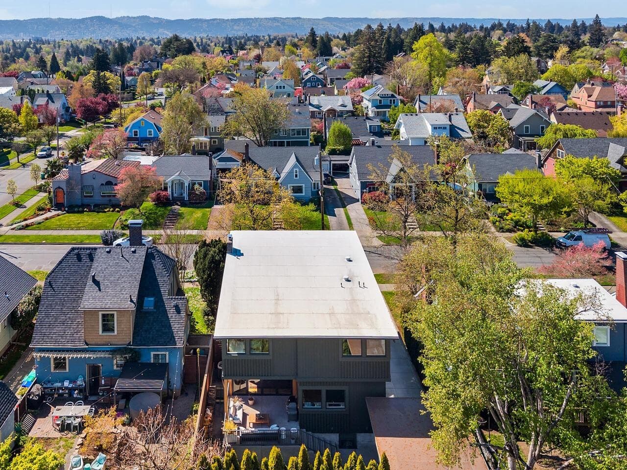 Aerial view of a suburban neighborhood with colorful houses, tree-lined streets, and yards. The image shows homes with pitched and flat roofs, surrounded by green trees and gardens on a clear, sunny day.