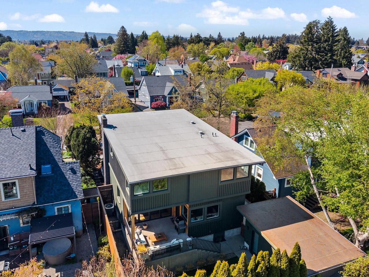 Aerial view of a modern two-story house with a flat roof, surrounded by trees and neighboring homes in a suburban neighborhood on a clear, sunny day.