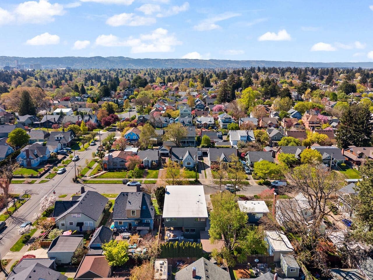 Aerial view of a suburban neighborhood with tree-lined streets, single-family homes, and greenery under a blue sky with scattered clouds. Hills are visible in the distance.