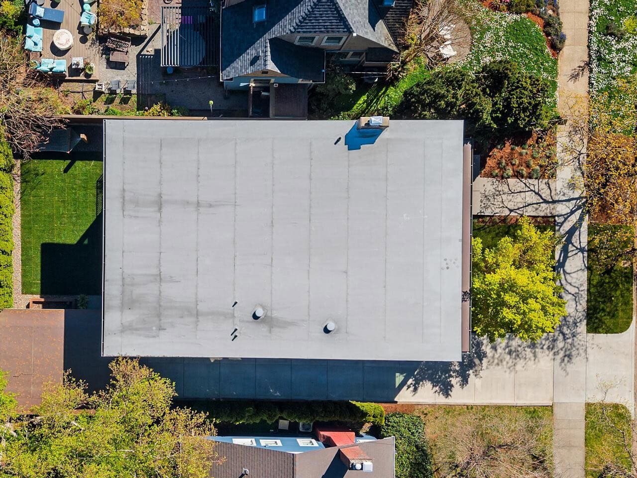 Aerial view of a rectangular flat-roofed building surrounded by green trees, yards, and neighboring houses, with a driveway and sidewalk visible along one side.