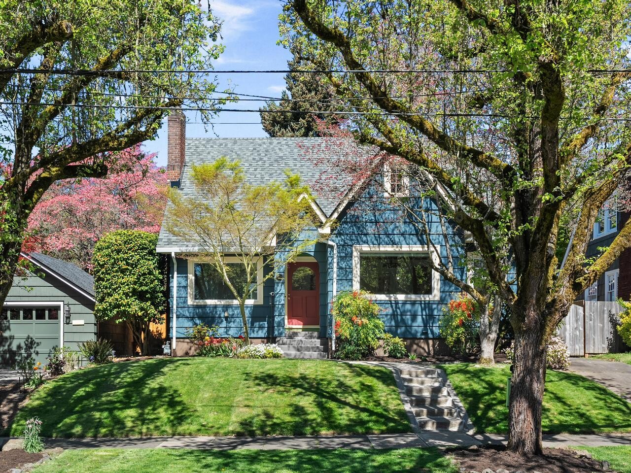 A charming blue house with a red front door sits behind a lush green lawn, surrounded by large trees and colorful shrubs, on a sunny day. Concrete steps lead up from the sidewalk to the front porch.