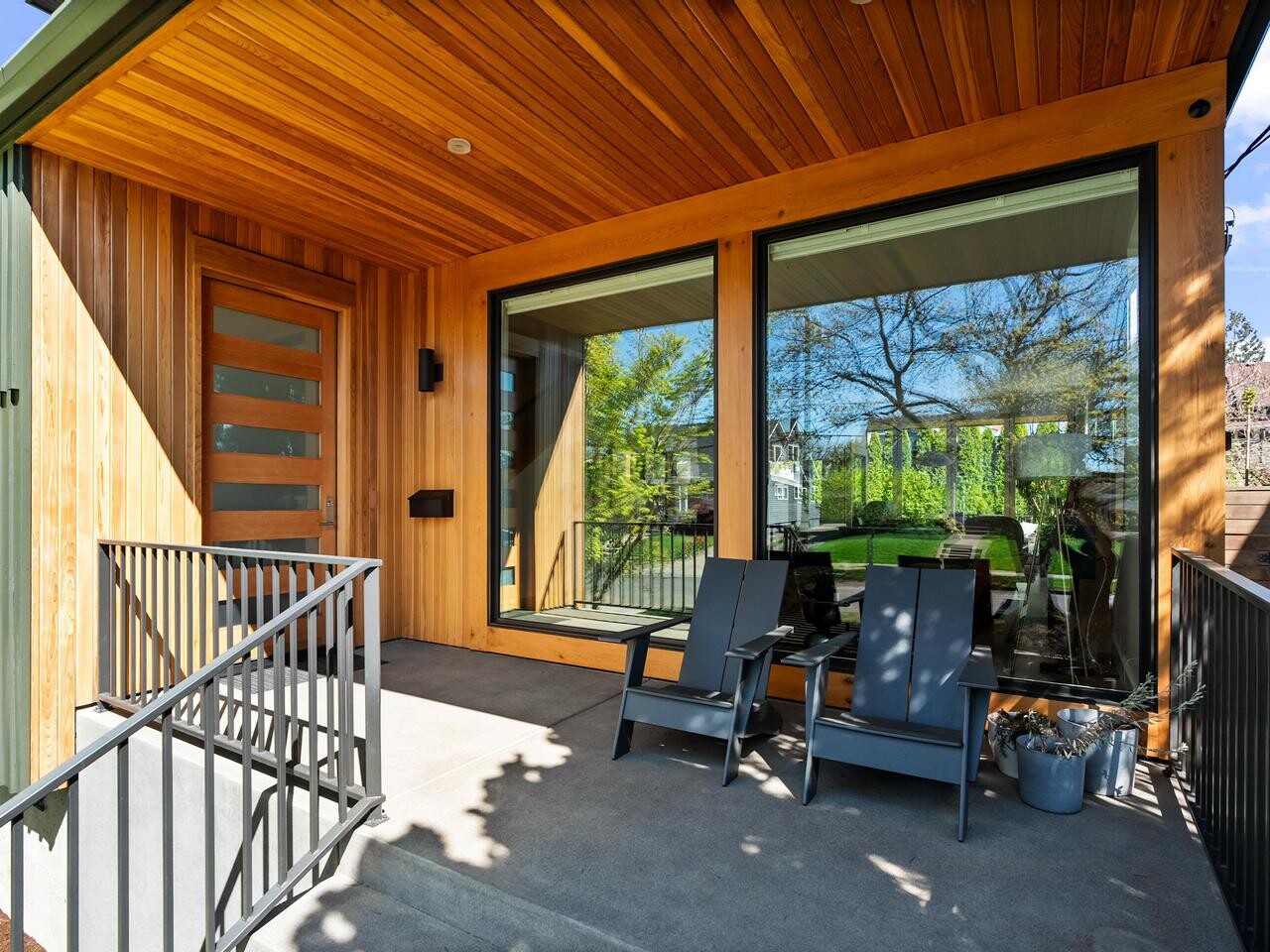 A modern wooden front porch with a slatted ceiling, large windows, a glass-panel door, two black chairs, a small table, metal railings, and a view of a green backyard with trees and houses.