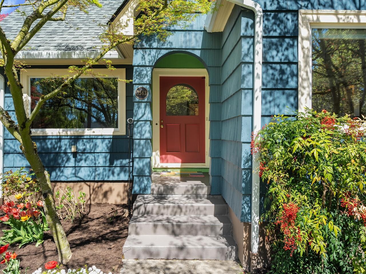 A blue house with white trim and a red front door, partially shaded by tree branches. Colorful flowers and shrubs grow along the walkway and near the entrance. Sunlight filters through the leaves.