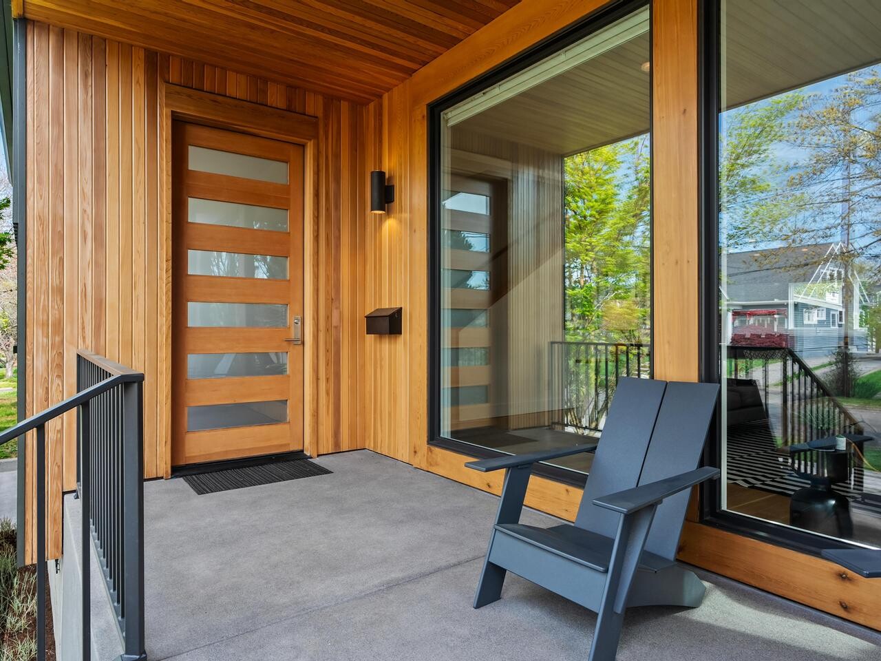 Modern front porch with light wood siding, a large window, a wooden door with horizontal glass panels, black railing, and a gray Adirondack chair facing the street. Trees and houses are visible in the background.