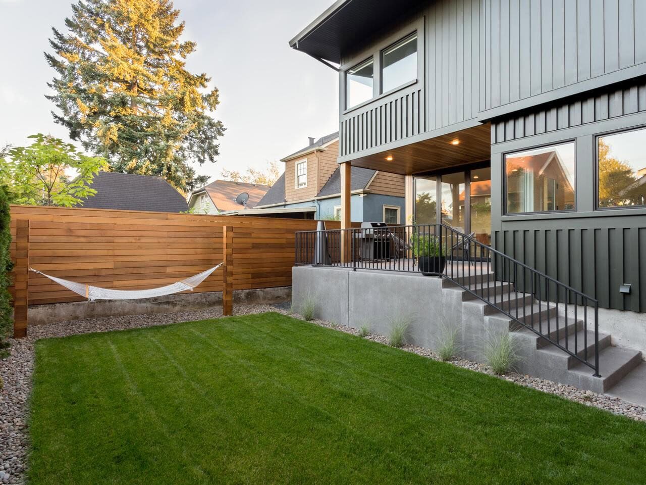 A modern backyard with a green lawn, a white hammock hanging between wooden posts, a wooden privacy fence, and a covered patio area attached to a gray house with stairs leading down to the yard.