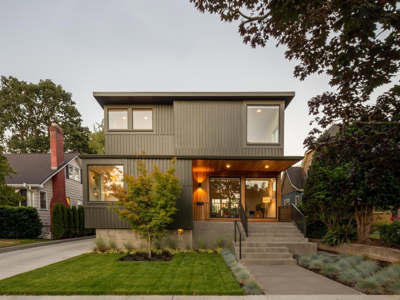 Modern two-story house with large windows, gray metal siding, a covered entryway, and landscaped front yard with grass, small tree, and concrete steps leading to the front door.