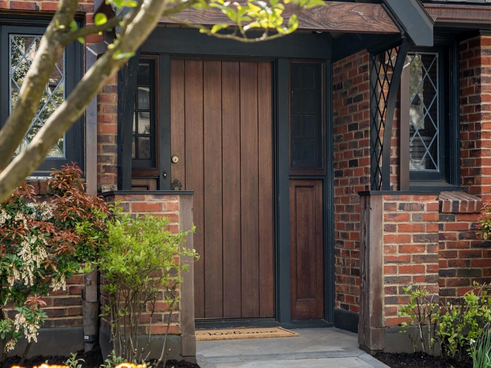 A wooden front door of a brick house is surrounded by greenery and flowering plants. Yellow and orange tulips bloom in the foreground, with a tree branch partially framing the walkway leading to the entrance.
