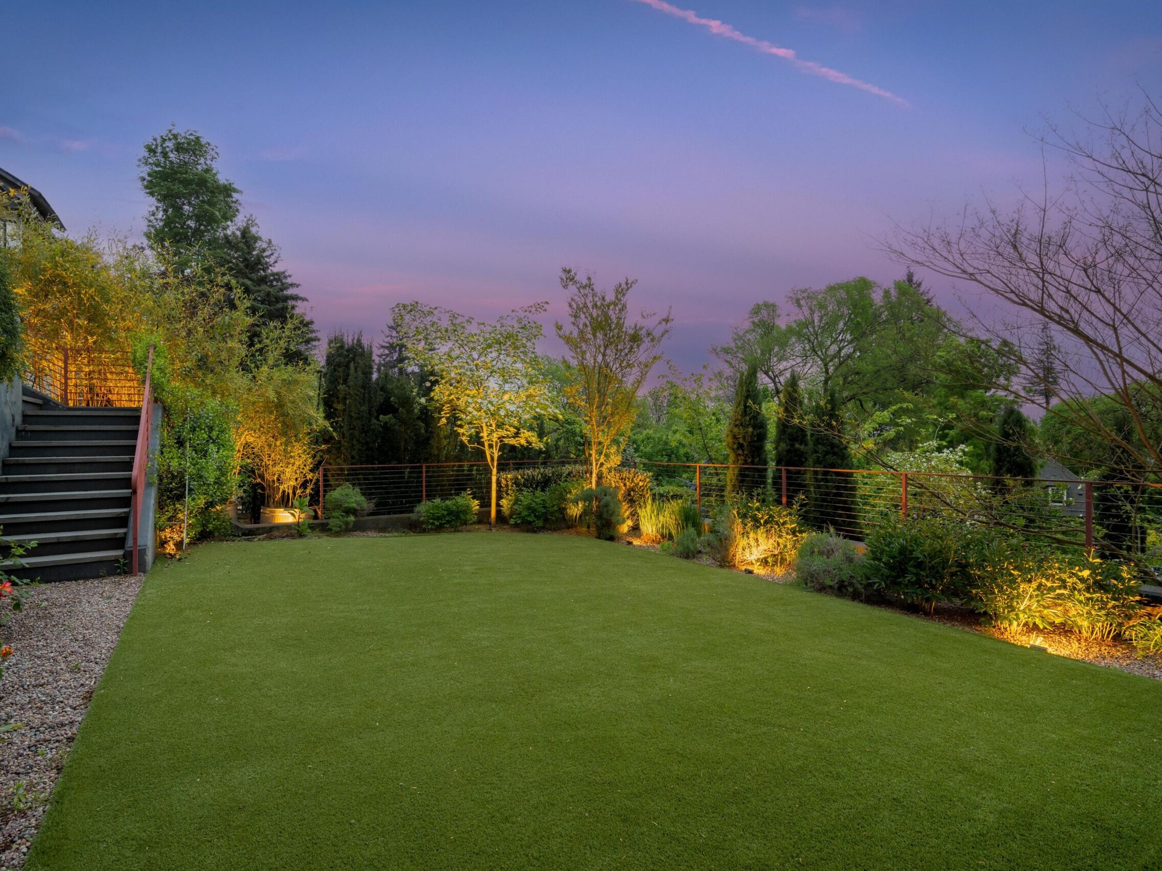 A landscaped backyard at dusk features a manicured lawn, illuminated trees and shrubs, gravel pathways, and steps leading up to a patio, with a purple sky in the background.