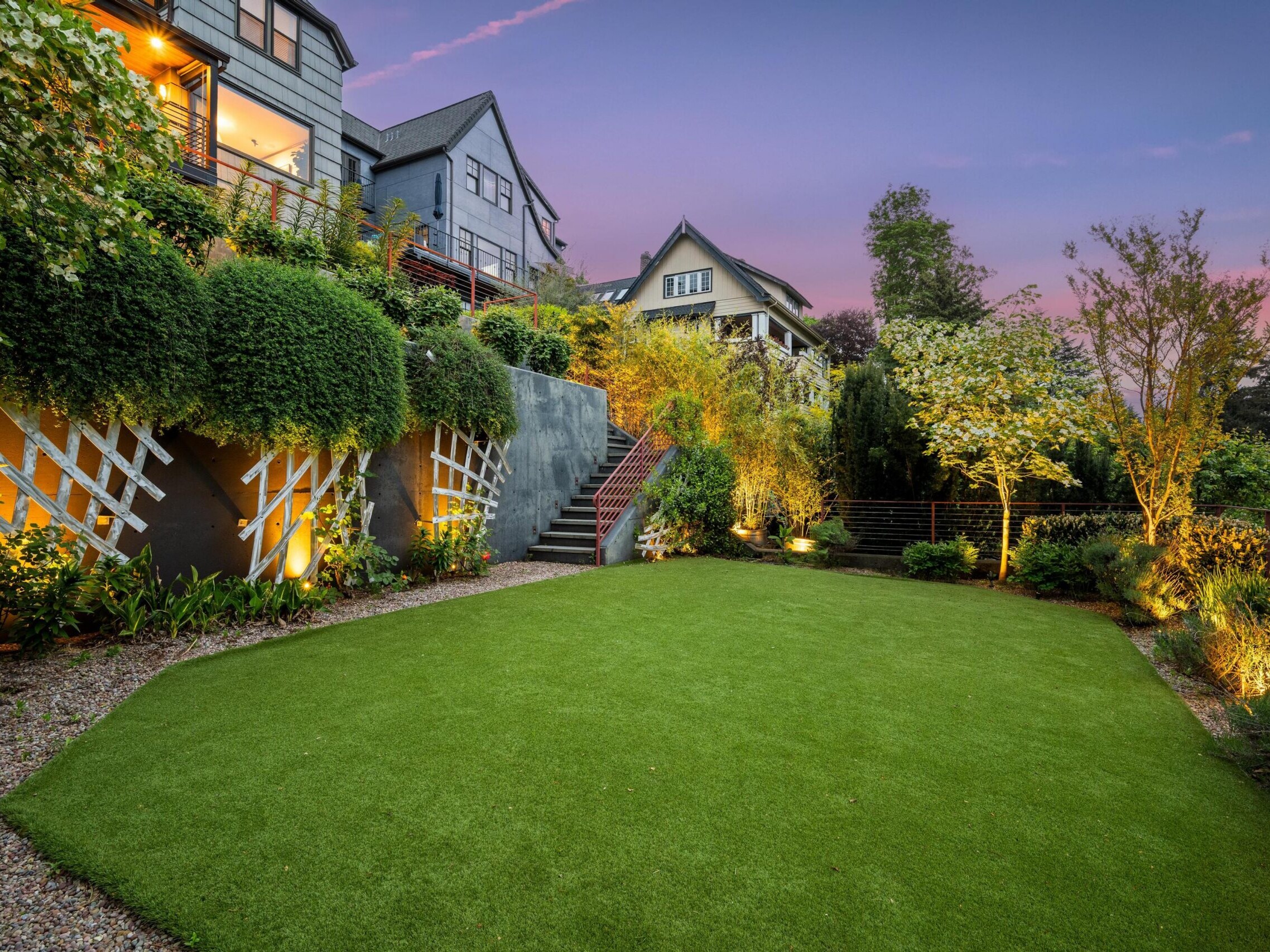 A landscaped backyard at sunset features a manicured lawn, lush greenery, decorative trellis panels on a retaining wall, and residential houses in the background with warm outdoor lighting.