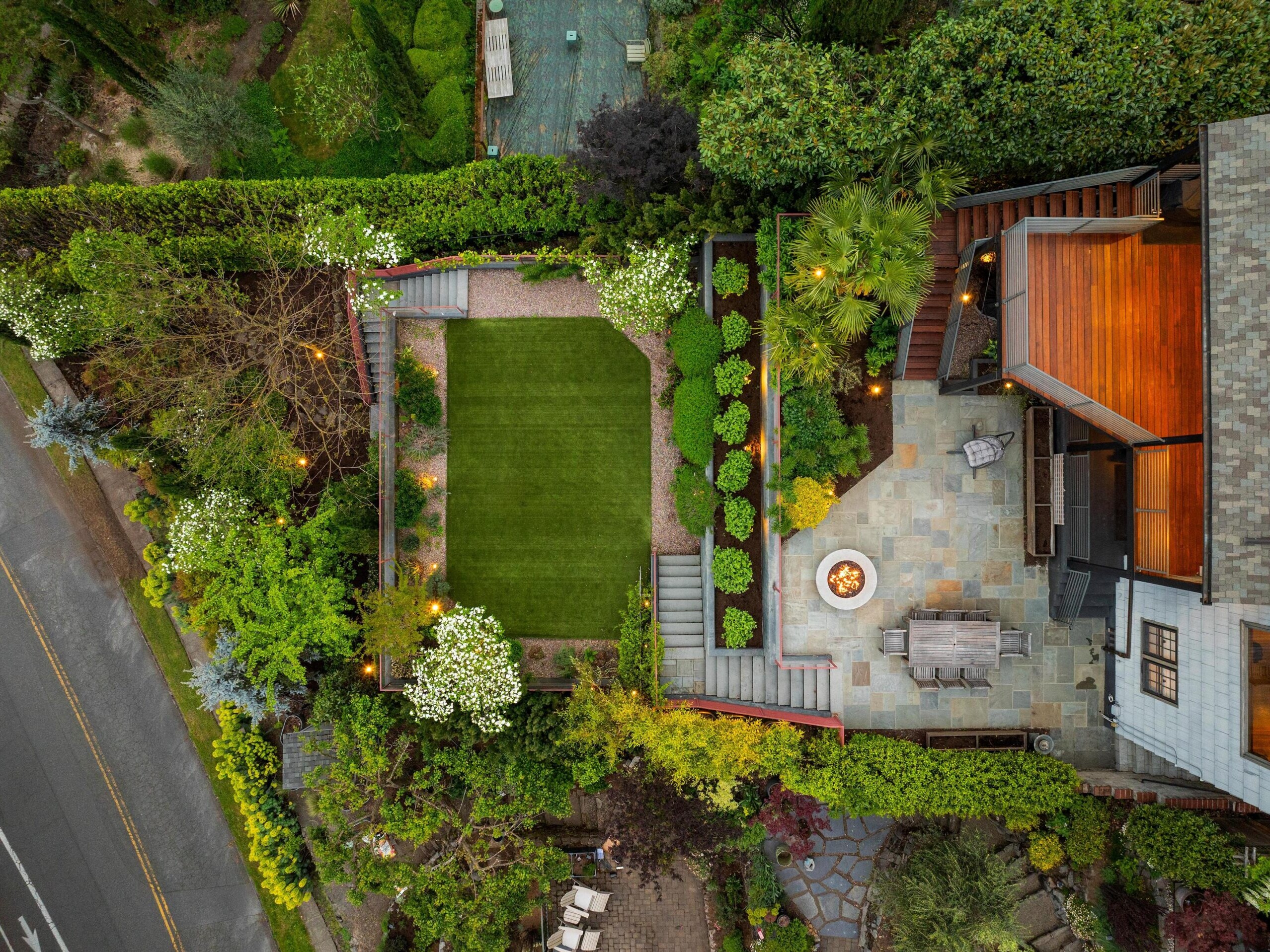 Aerial view of a landscaped backyard featuring a rectangular lawn, lush greenery, a stone patio with seating around a fire pit, and a wooden deck attached to a house. Pathways and trees border the yard.