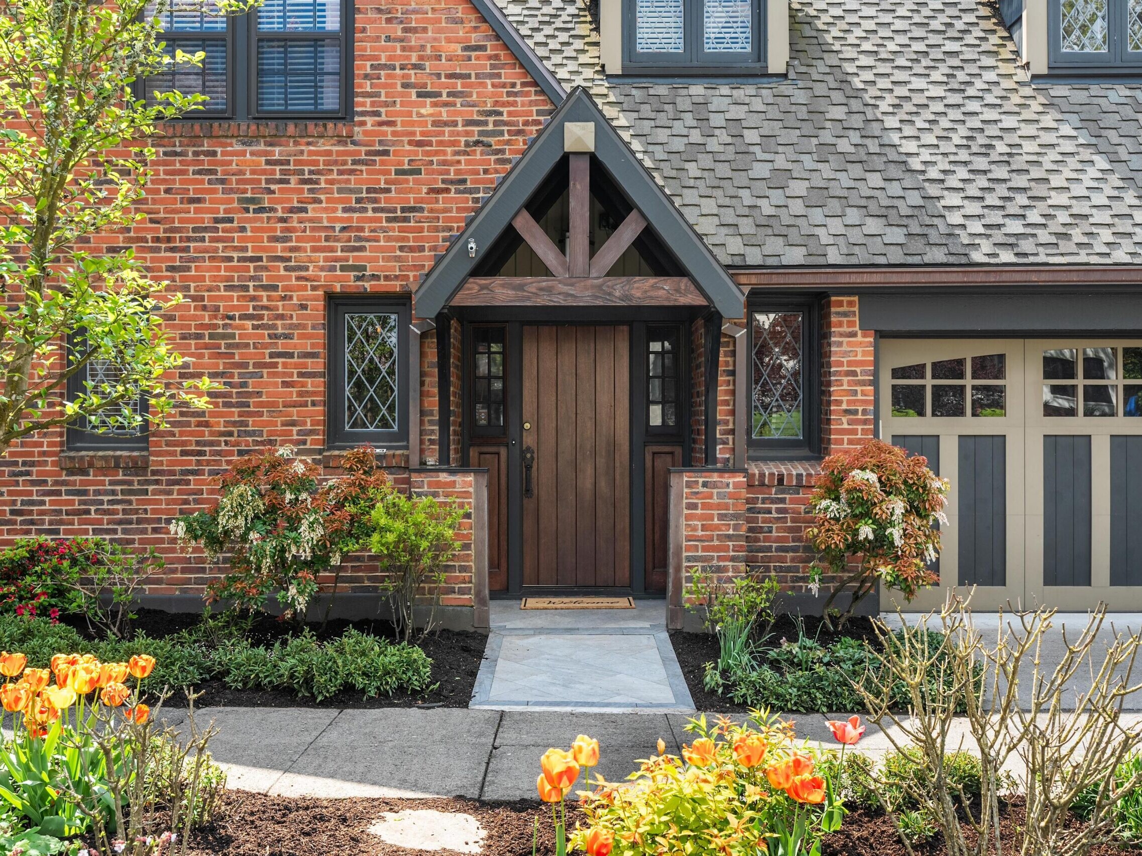 A brick house with a wooden front door, black trim, and diamond-patterned windows. Tulips bloom in the landscaped garden, and a two-tone garage door is visible on the right.